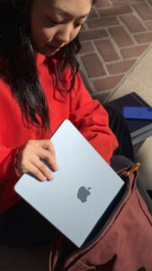 "A student in a red hoodie sliding a sleek blue Apple MacBook Air into a backpack while sitting outdoors."