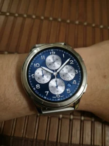 Amazfit Active 3 Premium smartwatch worn on wrist, showing a classic blue analog chronograph-style watch face with multiple sub-dials and a date window, against a wooden background.