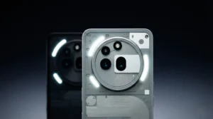 White Nothing Phone 4a shown from the rear, centered against a dark gradient background. The image focuses on the large circular camera module with multiple lenses and a rectangular sensor cutout, surrounded by illuminated white Glyph LED strips. A black variant appears softly blurred in the background.